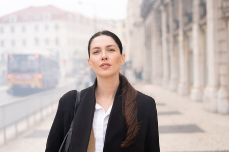 Portrait of businesswoman in black coat with long brown hair standing on sidewalk. Woman has confident expression with focused gaze. Soft light highlights her face against blurred background of buildingsの写真素材