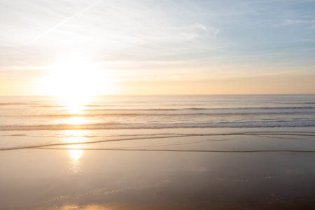 Peaceful scene of setting sun casting warm glow over sea waves and sandy shore. Tranquil atmosphere with soft colors and gentle water movement at sunset beachの写真素材