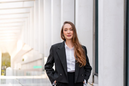 Businesswoman in black suit and white shirt standing with confident smile in modern city area. Young...