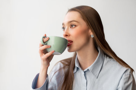 Young woman in shirt drinking coffee from cup while gazing away indoors with relaxed expression and natural lightの写真素材