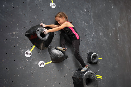 Determined girl climbing artificial wall at bouldering center. Brave and strong female child tackling mock wall, full body side view of sporty girl in actionの写真素材