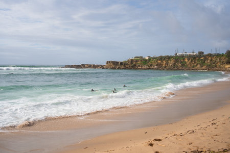 Group of surfers floating in sea near rocky shoreline. Peaceful beach with light waves and cloudy sky. Coastal scene capturing nature and outdoor water activityの写真素材