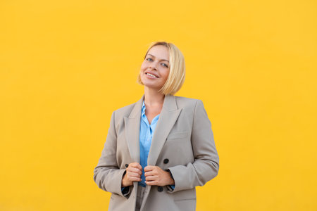 Young businesswoman with short blond hair wearing beige blazer holding blazer while smiling confidently against bright yellow backgroundの写真素材