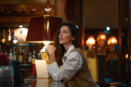 Young woman in white shirt and vest sitting at bar counter looking thoughtful. Warm light from vintage lamp creates cozy and relaxed atmosphere in luxury settingの写真素材