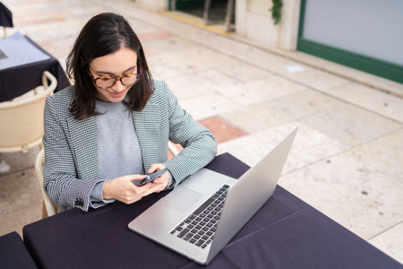 Brunette woman in casual clothes working with laptop using mobile phone at cafe. Freelancer using smartphone and laptop sitting outdoor cafeの写真素材