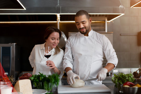 Handsome young African chef is cooking together with Caucasian girlfriend in the kitchen using red wine ingredient. A cook teaches a girl how to cook. Man and woman cooking in professional kitchen.の写真素材