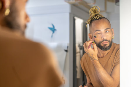 Black Brazilian gay man confidently applying makeup to his face as he gazes into mirror. Beauty and strength of self-expression and celebrates diversity of gender and sexualityの写真素材