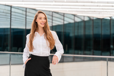 Smiling businesswoman in white shirt and black pants standing with hand in pocket near modern glass office building facade, confident professional posing in contemporary urban settingの写真素材