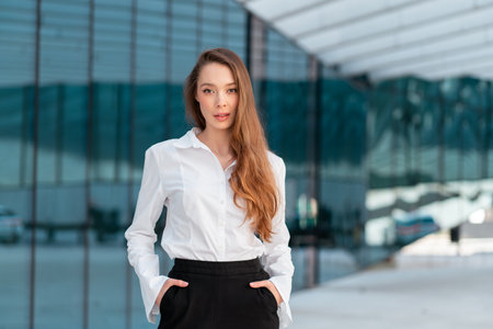 Confident businesswoman in white shirt and black pants standing with hands in pockets near glass building, reflecting modern professionalism and charmの写真素材