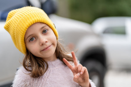 Young girl wearing yellow cap making a peace sign while smiling at camera. She looks calm and...