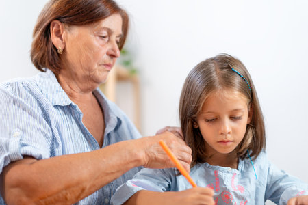 Grandmother watching girl drawing with colored pencil at home showing encouragement and admiration while child focuses on creative activityの写真素材