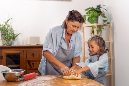 Grandmother guiding granddaughter to roll dough on kitchen table while baking together at home and enjoying fun family cooking activityの写真素材