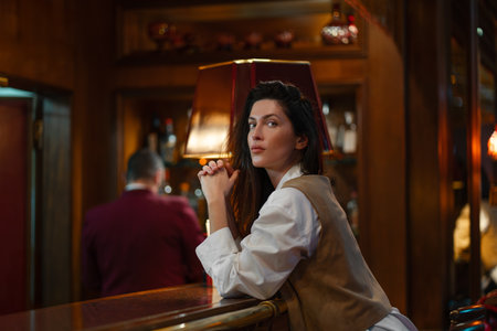 Stylish woman in vest and white shirt leaning on bar counter in dim lounge with serious and confident expression enjoying nightlife atmosphereの写真素材
