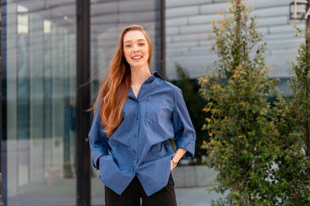 Smiling businesswoman in blue shirt standing near modern glass office building with confident and positive expression during daytimeの写真素材