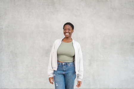 Smiling young black woman standing against gray wall wearing trendy casual clothes. Friendly expression with relaxed mood and modern everyday styleの写真素材