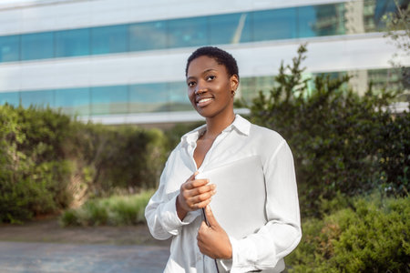 Confident young businesswoman in white shirt with laptop standing near modern office building. Thoughtful female professional is smiling and looking awayの写真素材