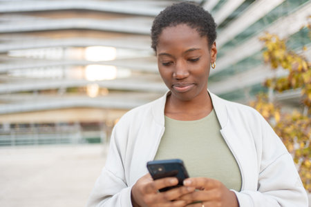 Young businesswoman in smart casuals using smartphone while standing near modern office building. Focused expression and digital communication conceptの写真素材