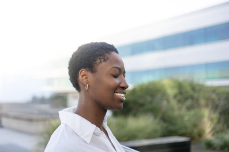 Side view of smiling young black businesswoman outside modern office building. Female professional is wearing white shirt with eyes closed during daytimeの写真素材