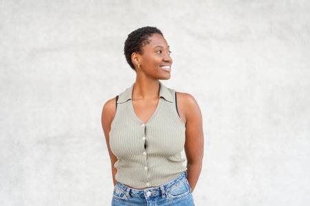 Smiling young woman looking away while standing against gray wall. Thoughtful mood with relaxed posture and modern casual styleの写真素材