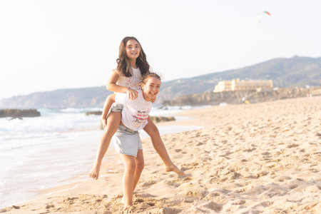 Cheerful girls playing piggyback ride on sandy beach near sea smiling and enjoying fun time during sunny dayの写真素材