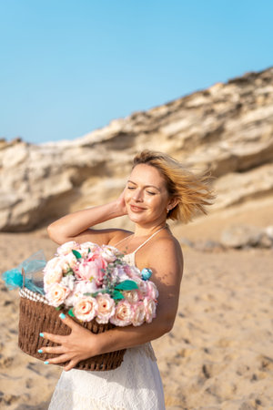 Smiling young woman with windswept hair holding basket with pink flowers on sandy beach enjoying summer vacation near rockの写真素材