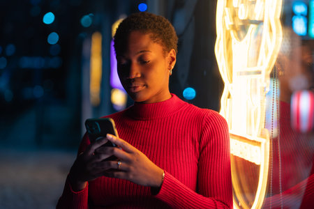 Young black woman using smartphone beside glowing neon lights on city street at night. Female in red sweater text messaging on mobile phoneの写真素材