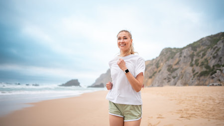 Smiling woman jogging along sandy beach in morning. Athletic female in white top and green shorts running by ocean shore, enjoying nature and fitnessの写真素材