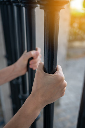 Closeup of hands of anonymous person gripping metal gate bars in sunlight during sunsetの写真素材