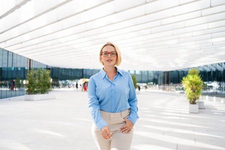 Young businesswoman with short blond hair wearing blue shirt standing confidently in front of modern office building during daytimeの写真素材