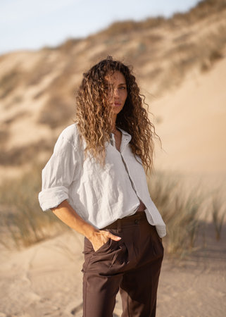 Portrait of trendy woman in white shirt and brown pants standing on sandy beach dune with hands in pockets. Confident lady with curly long hair looking at cameraの写真素材