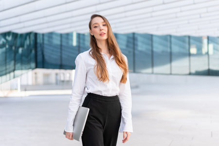 Confident businesswoman in white shirt and black pants standing with closed laptop near modern office building. Professional confidence and corporate work conceptの写真素材