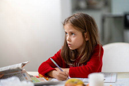 Girl in red pajamas sitting at table and doing homework at home. Thoughtful expression during learning time with focus and concentration conceptの写真素材