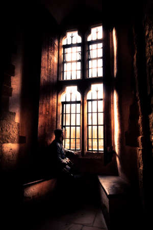 Rear view at silhouette of man sitting in the darkness looking through old bright window with incoming rays of light, dreaming, resting, thinking deeply while waiting quietly and day goes by.の写真素材