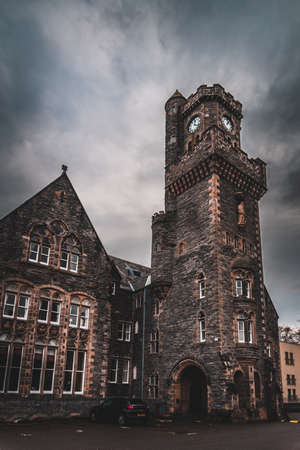 FORT AUGUSTUS, SCOTLAND, DECEMBER 17, 2018: The Abbey Highland Club clock tower, full of mold and lichen in its stone walls, under heavy cloudscape with mountains in the background, aside Loch Nessのeditorial素材