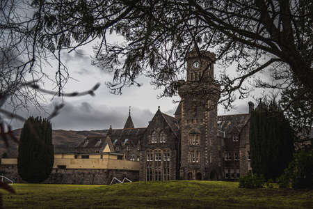 FORT AUGUSTUS, SCOTLAND, DECEMBER 17, 2018: The Abbey Highland Club clock tower, full of mold and lichen in its stone walls, under heavy cloudscape with mountains in the background, aside Loch Nessのeditorial素材