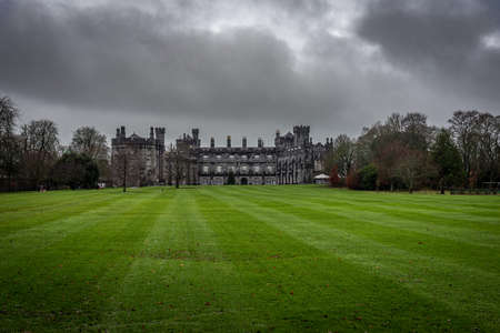 KILKENNY, IRELAND, DECEMBER 23, 2018: Kilkenny Castle seen from the huge garden on a dramatic cloudy day with dry leaves over the green grass while people run beside it.のeditorial素材