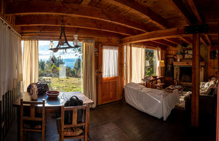 BARILOCHE, ARGENTINA, JUNE 18, 2019: Panoramic view of interior of a cozy cabin with comfortable couches, a fireplace, illuminated by sunlight from the window that sees to the forest and lake.のeditorial素材