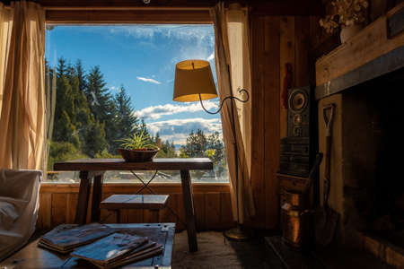 BARILOCHE, ARGENTINA, JUNE 18, 2019: Interior of a cozy and relaxing cabin with comfortable couches around the fireplace, illuminated by the sunlight from the window that sees to the forest and lakeのeditorial素材