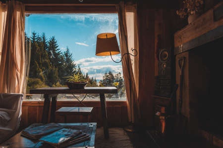 BARILOCHE, ARGENTINA, JUNE 18, 2019: Interior of a cozy and relaxing cabin with comfortable couches around the fireplace, illuminated by the sunlight from the window that sees to the forest and lakeのeditorial素材