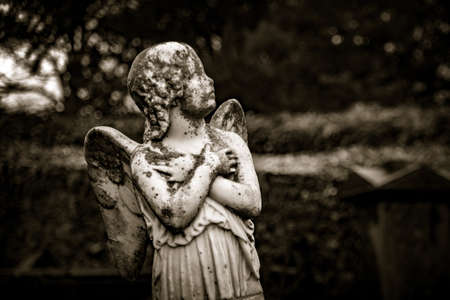 KILKENNY, IRELAND, DECEMBER 23, 2018: Sculpture of an old creepy cherub angel in the middle of a graveyard, full of lichen and mold, holding its chest and heart while looking at the cloudy sky. Concept of death, desolation and afterlifeのeditorial素材