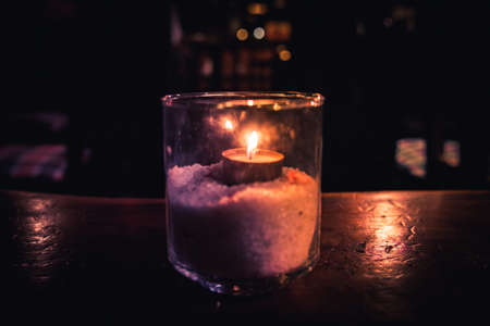 Isolated tealight inside a glass illuminating a dark scene and reflections on a wooden table and a nice bokeh background.の写真素材