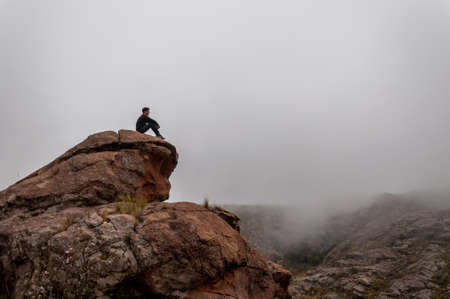 Young man sitting at the mountain peak staring at the dense fog along the mountainの写真素材