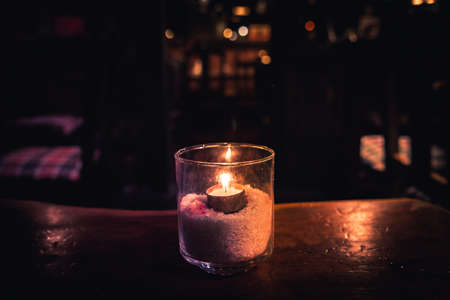 Isolated tealight inside a glass illuminating a dark scene and reflections on a wooden table and a nice bokeh backgroundの写真素材