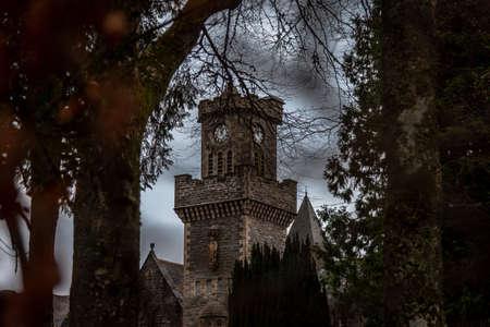 FORT AUGUSTUS, SCOTLAND, DECEMBER 17, 2018: The Abbey Highland Club clock tower, across leaves of trees, under heavy cloudscape with mountains in the background, aside Loch Ness.の写真素材