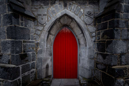 Scary pointy red wooden door in an old and wet stone wall building with cross, skull and bones at both sides. Concept mystery, death and danger.の写真素材