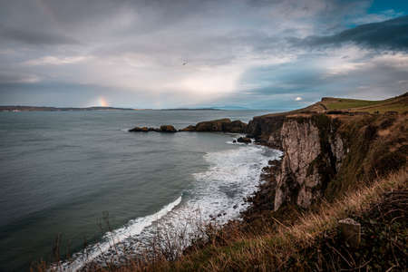 Rainbow shining between rainy clouds, sea and cliffs near Ballintoy in Northern Ireland, while a bird flies freely in the coast.の写真素材