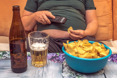 A fat man suffers from overweight and eats chips, drinks alcohol and looks in the phone. Concept: Healthcareの写真素材