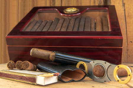 Time to enjoy! Quality cigars and other accessories such as cigar cutters, matches and cigar cases on a wooden table. In the background a humidor in the front of a wooden wall. Concentrate on the cigars. Cの写真素材