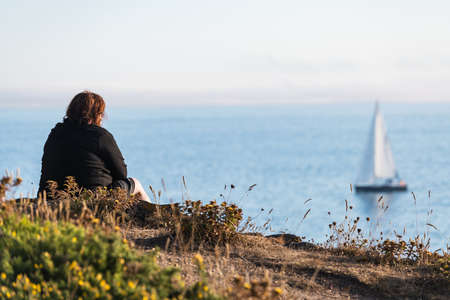 A lonely middle-age woman watches a boat sail away from Ons Island in the Ria de Pontevedra in Galicia at dusk.の写真素材