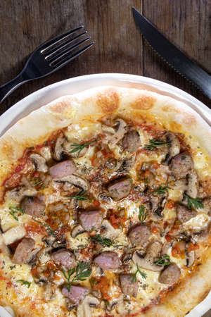 Ready-to-eat pizza with red sauce, mozzarella cheese, longaniza, mushroom, red onion and cilantro on a white plate on a wooden background with a fork and a knifeの写真素材